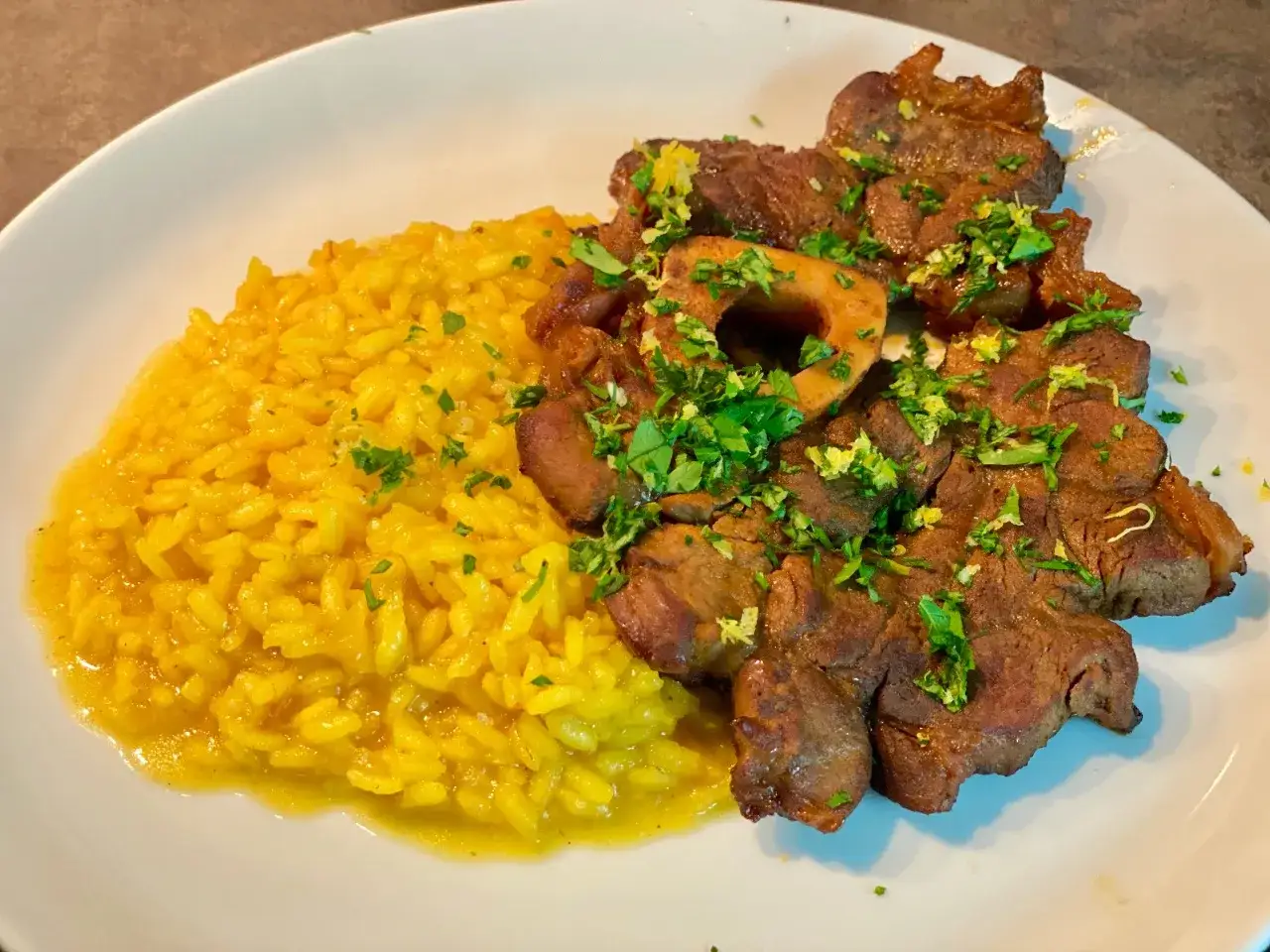 Ossobuco alla milanese succulento su un letto di risotto giallo brillante, guarnito con prezzemolo fresco e scorza di limone.