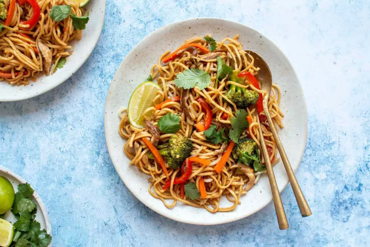 Spaghetti di soia con verdure colorate, broccoli, peperoni e carote, guarniti con coriandolo fresco e spicchi di lime.
