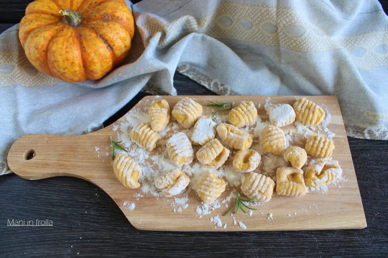 Gnocchi di zucca fatti in casa, spolverati di farina, su un tagliere di legno, con una zucca arancione sullo sfondo.