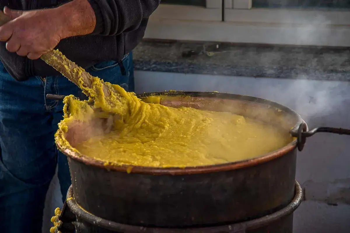 Un uomo mescola con un mestolo di legno una polenta fumante preparata con farina di mais in un grande paiolo di rame.