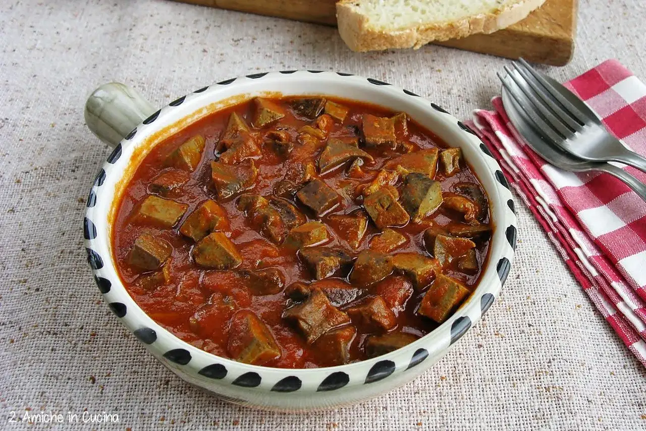 Piatto fumante di coratella di agnello in umido, servita in una ciotola decorata. Accanto, pane fresco e posate.