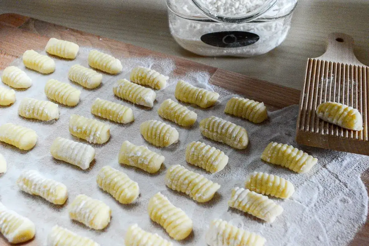 Gnocchi di patate freschi, pronti per essere cotti, disposti su un tagliere infarinato. Accanto, un rigagnocchi e un barattolo di farina.