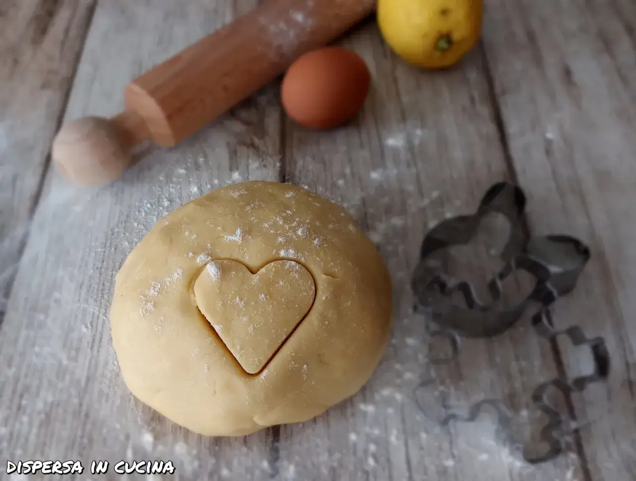 Impasto per pasta frolla senza burro con un cuore inciso, mattarello, uovo e limone pronti per biscotti fatti in casa.
