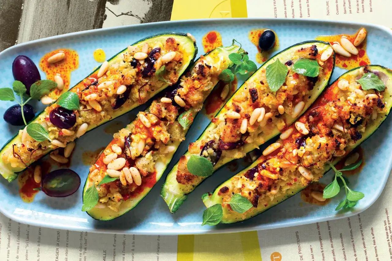 Zucchine ripiene vegetariane con pinoli, olive e erbe aromatiche, servite su un piatto azzurro con un filo d'olio piccante.
