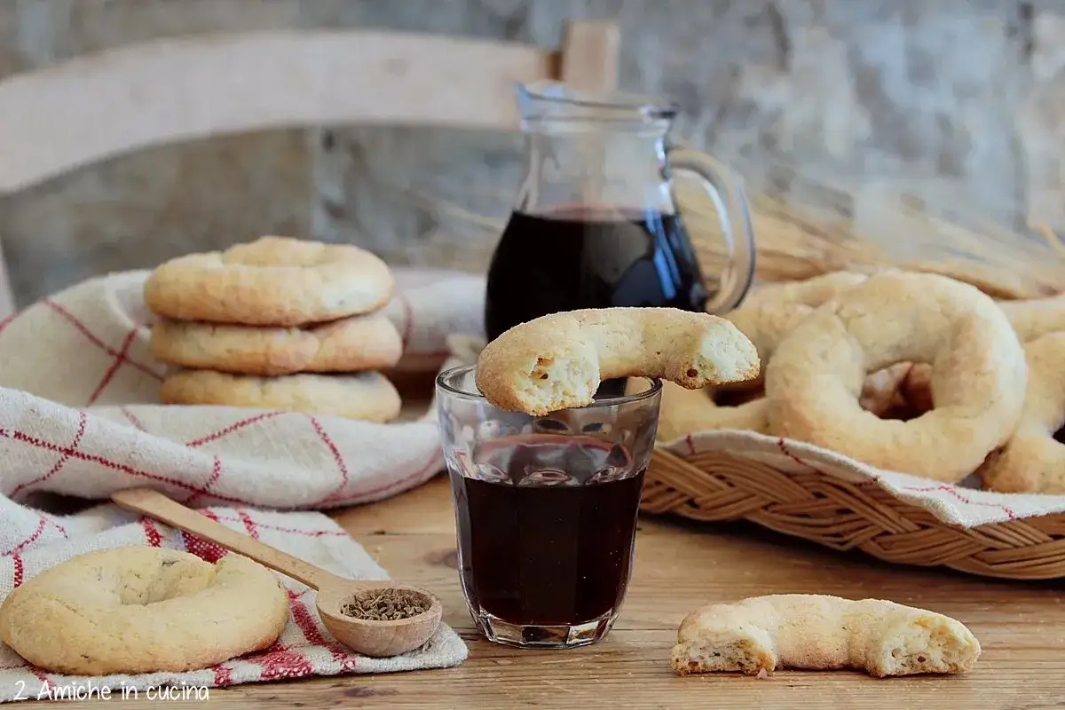 Ciambelline al vino dorate, una su un bicchiere di vino rosso, su un tavolo di legno con una brocca e una spiga.