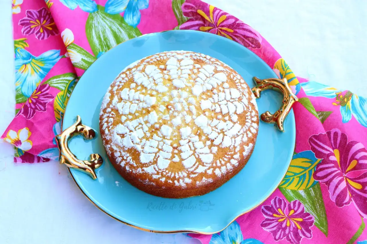 Torta senza lievito, soffice e dorata, decorata con zucchero a velo, su un piatto azzurro con manici dorati, adagiata su un tovagliolo fiorato.