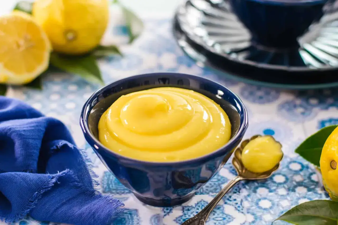 Una ciotola blu piena di vellutata crema pasticcera al limone, con limoni freschi e un tovagliolo blu sullo sfondo.
