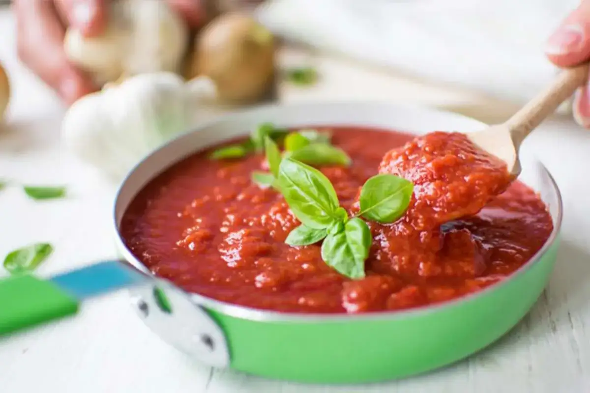 Un cucchiaio di legno raccoglie un ricco sugo al pomodoro, guarnito con basilico fresco, da una padella verde. Sullo sfondo, aglio e cipolle.
