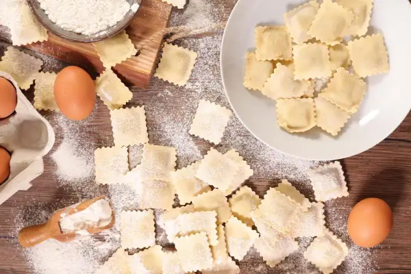 Ravioli fatti in casa sparsi su un tavolo di legno, con farina, uova e una ciotola di farina.