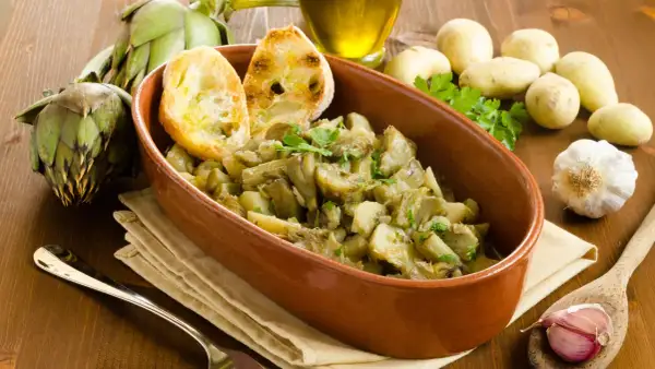 Un saporito piatto di carciofi in umido con patate, guarnito con pane tostato e prezzemolo fresco. Ingredienti freschi come carciofi, aglio e patate completano la scena.