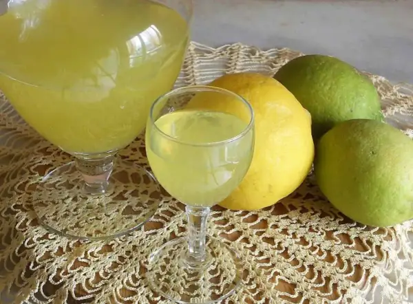 Limoncello ricetta originale Amalfi: bicchieri di liquore giallo brillante e limoni freschi, alcuni verdi, altri gialli, su un centrino di pizzo.