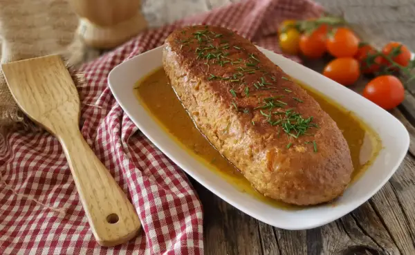 Un succulento polpettone in padella, dorato e profumato, adagiato su un letto di salsa e guarnito con erba cipollina. Accanto, una spatola in legno e pomodorini freschi.