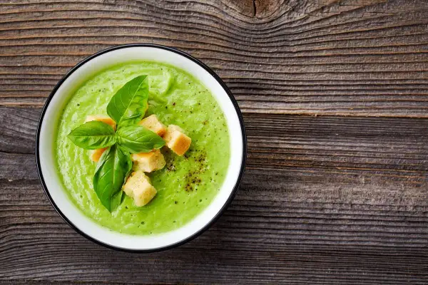 Un cremoso passato di verdure verde brillante, guarnito con crostini dorati e foglie di basilico fresco, su un tavolo di legno rustico.
