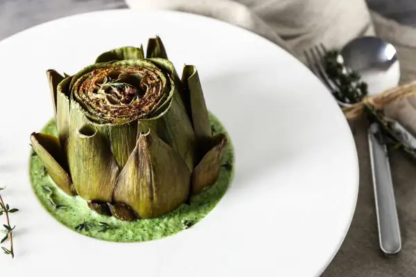 Carciofo intero cotto, adagiato su una vellutata crema di carciofi, guarnito con erbe aromatiche.