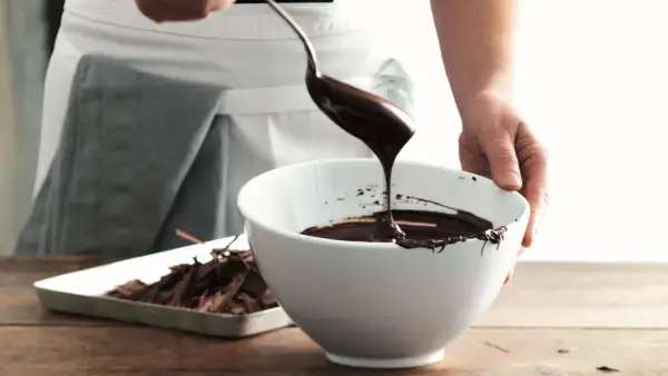 Mani che versano cioccolato fuso da un cucchiaio in una ciotola bianca, mostrando come sciogliere il cioccolato per dolci deliziosi.