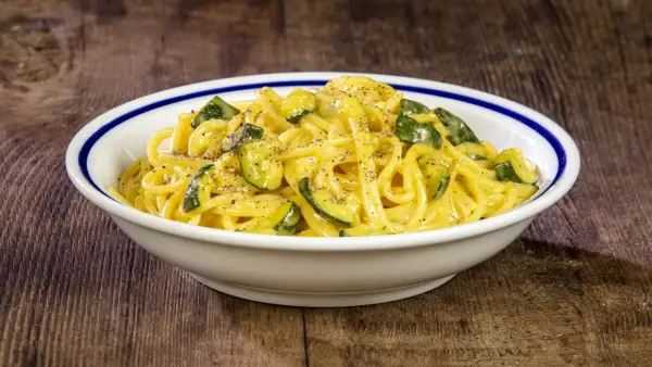 Spaghetti with zucchini and creamy sauce in a white bowl with blue trim on a wooden table.