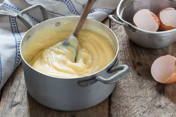 Crema pasticcera veloce in pentola, con cucchiaio di legno che mescola. Guscio d'uovo accanto, pronto per un dolce.