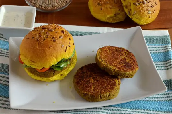 Hamburger di lenticchie dorato con semi, servito su un piatto bianco con due polpette aggiuntive e una ciotola di salsa.