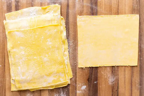 Fogli di pasta gialla, pronti per diventare deliziose lasagne fatte in casa. Un foglio singolo è posato a destra, mentre una pila di altri è a sinistra.