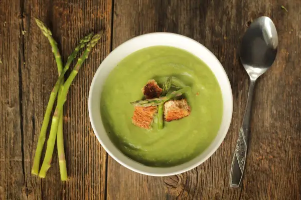 Crema di asparagi vellutata, guarnita con crostini e asparagi freschi, su un tavolo di legno rustico.