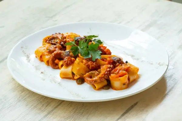 Piatto di paccheri con ragù di polpo, pomodorini e capperi, guarnito con prezzemolo fresco.