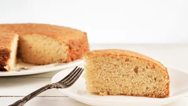 Una fetta di torta senza lievito, soffice e dorata, attende su un piatto bianco con una forchetta antica.