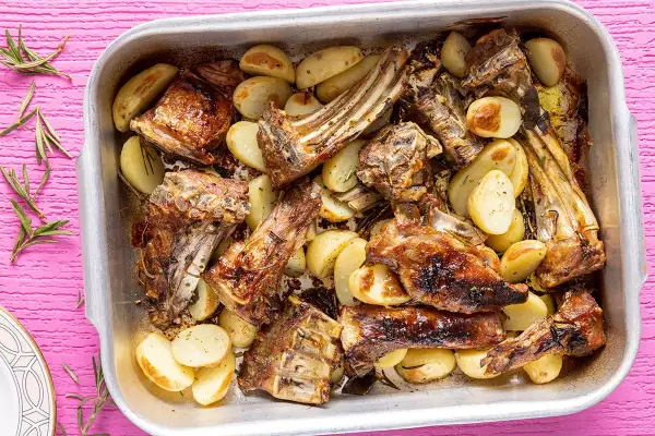 Succulento abbacchio al forno con patate dorate, profumato al rosmarino, servito in una teglia d'argento su un tavolo rosa acceso.