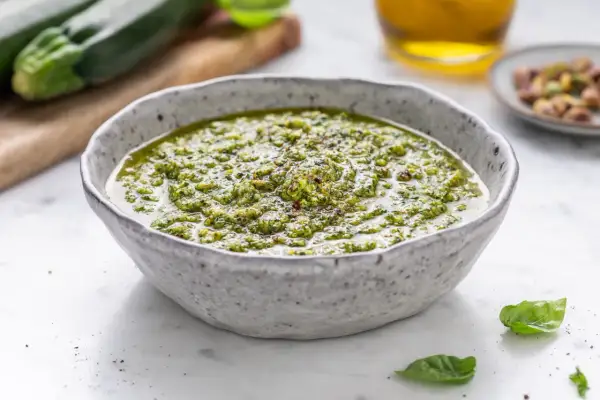 Pesto di zucchine cremoso in una ciotola, con zucchine fresche e pistacchi sullo sfondo.
