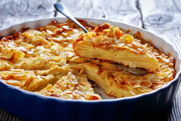 A slice of golden-brown apple cake with thin apple layers, topped with toasted almond slivers, is lifted from a blue baking dish.