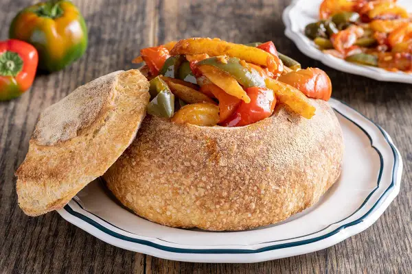Un pane scavato ripieno di un saporito stufato di pipi e patate, con una fetta di pane croccante a lato.