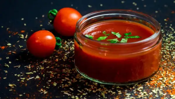 Un vasetto di sugo al pomodoro, guarnito con basilico fresco, accanto a pomodorini e spezie su sfondo scuro.