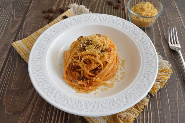 Un piatto di spaghetti con acciughe, pomodoro e pangrattato tostato, servito su un piatto bianco decorato, con un tovagliolo giallo e un bicchiere di pangrattato a lato.